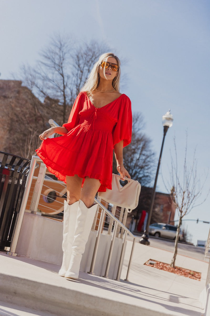 Full body view of model wearing the Rowyn Red Short Sleeve Dress which features red rayon fabric, three tiered body style, ruffle hem, mini length, red lining, smocked waist, v-neckline with tie closure and short puff sleeves.