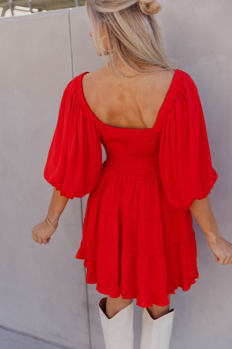 Back view of model wearing the Rowyn Red Short Sleeve Dress which features red rayon fabric, three tiered body style, ruffle hem, mini length, red lining, smocked waist, v-neckline with tie closure and short puff sleeves.