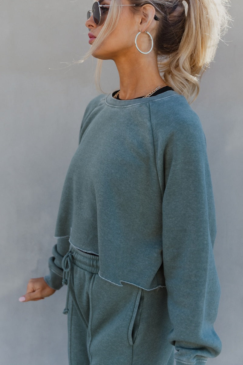 Side view of model wearing the Mia Grey Green Knit Long Sleeve Sweatshirt which features gray green knit fabric, cropped waist, raw hem, round neckline and long sleeves with cuffs.