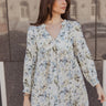 Front view of model wearing the Evelyn Blue Multi Floral Mini Dress that has white fabric with a floral pattern, mini length, two covered buttons, a round neckline with a plunge detail, and long sleeves with elastic wrists.