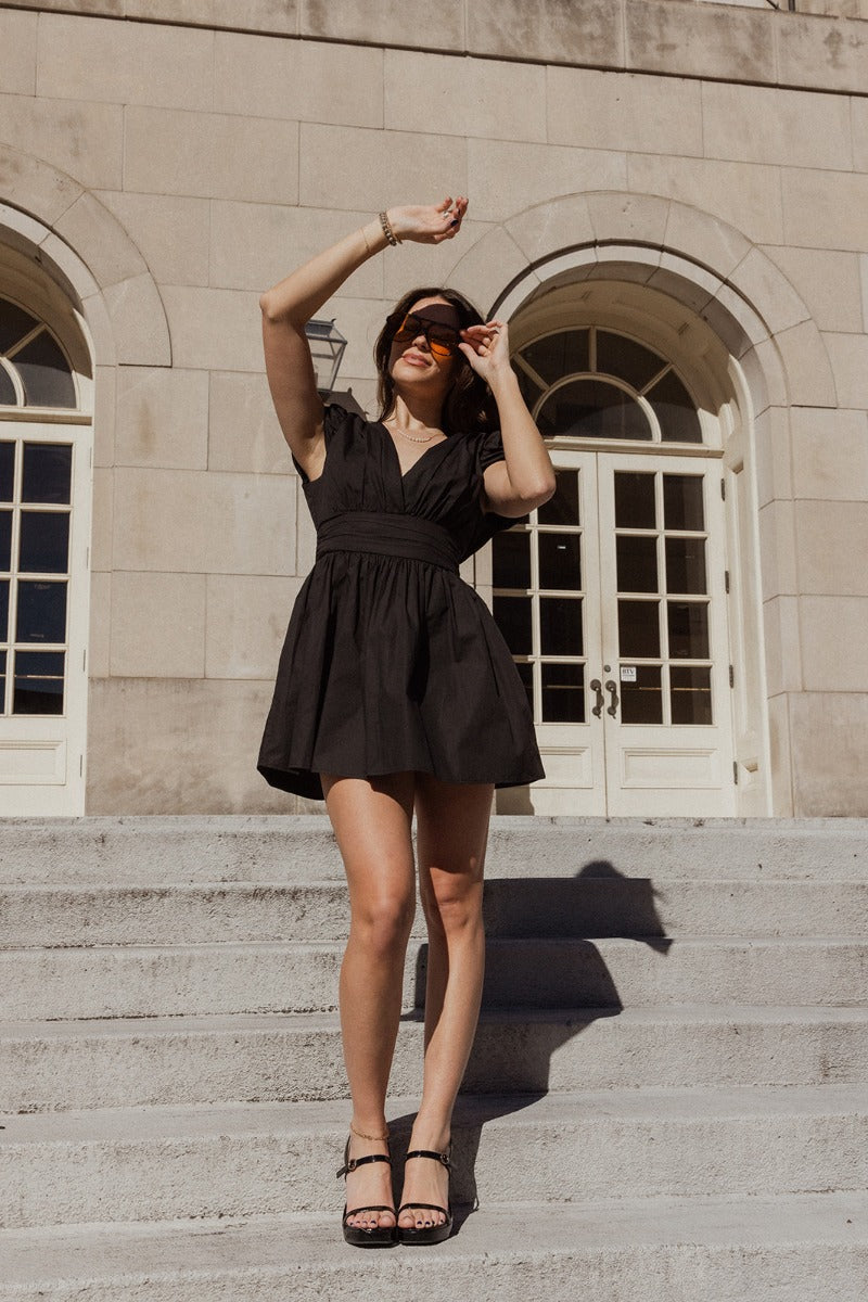 Full body view of model wearing the Jasmine Black Short Sleeve Mini Dress which features black lightweight fabric, black shorts lining, mini length, a folded detail waistband, an overlap plunge neckline, shorts sleeves, and a monochrome back zipper with a