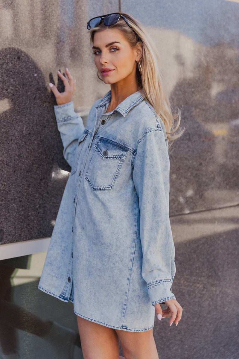 Frontal side view of model wearing the Jasmine Light Denim Wash Buttoned Long Sleeve Dress which features light wash denim fabric, button up closures, mini length, two front chest buttoned pockets, a collared neckline, and long sleeves with buttoned cuffs