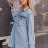 Frontal side view of model wearing the Jasmine Light Denim Wash Buttoned Long Sleeve Dress which features light wash denim fabric, button up closures, mini length, two front chest buttoned pockets, a collared neckline, and long sleeves with buttoned cuffs
