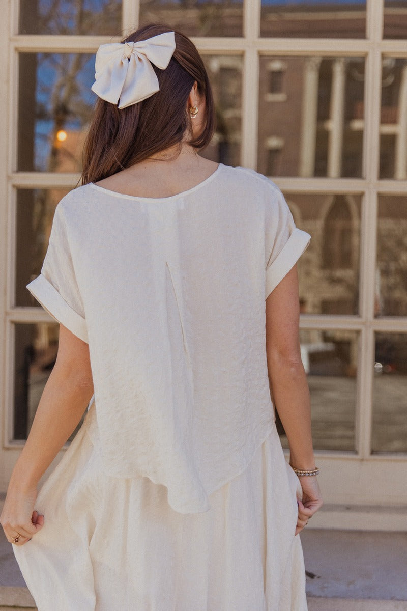 Back view of model wearing the Amelia Off White Short Sleeve Top which features ivory lightweight fabric with a monochrome pattern design, a scooped hem, a round neckline, and short sleeves with folded hem details.