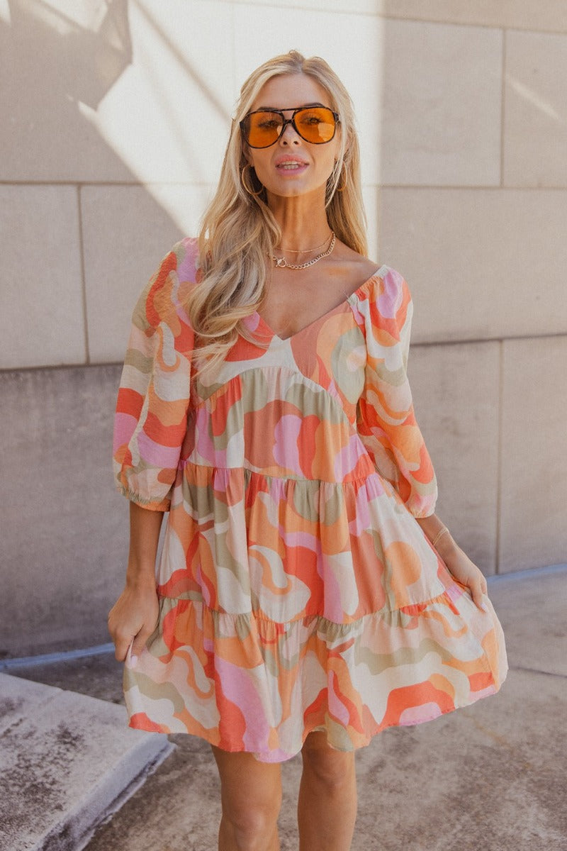 Front view of model wearing the Eloise Multi-Color Swirl Print Mini Dress that has blush, orange, peach, pink, beige, rust and sage fabric, a swirl pattern, two tiered body, mini length, a v-neck, a smocked back and half sleeves with elastic trim.

