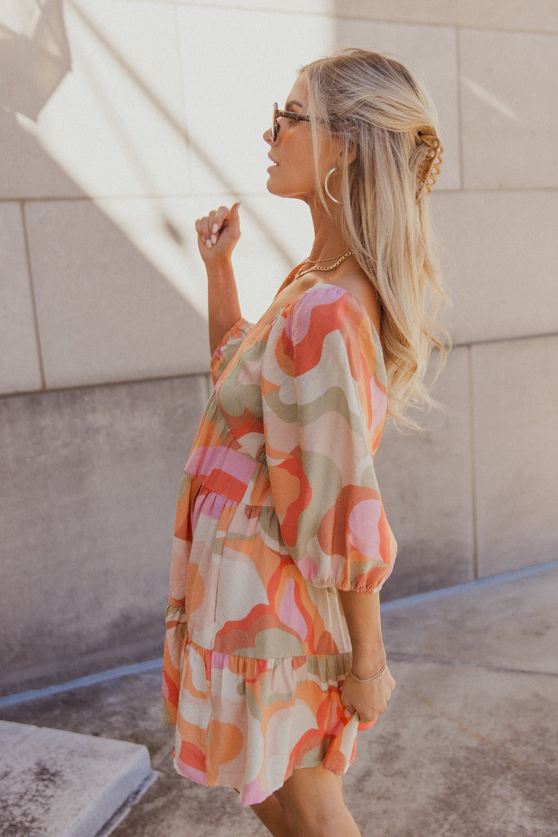 Side view of model wearing the Eloise Multi-Color Swirl Print Mini Dress that has blush, orange, peach, pink, beige, rust and sage fabric, a swirl pattern, two tiered body, mini length, a v-neck, a smocked back and half sleeves with elastic trim.
