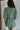 back view of model wearing the Celia Romper in Green that has green lightweight fabric, a bubble hem, a tie around the waist, a surplice neck, half puff sleeves with elastic trim, and a back zipper with a hook closure