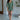 Full body front view of model wearing the Celia Romper in Green that has green lightweight fabric, a bubble hem, a tie around the waist, a surplice neck, half puff sleeves with elastic trim, and a back zipper with a hook closure