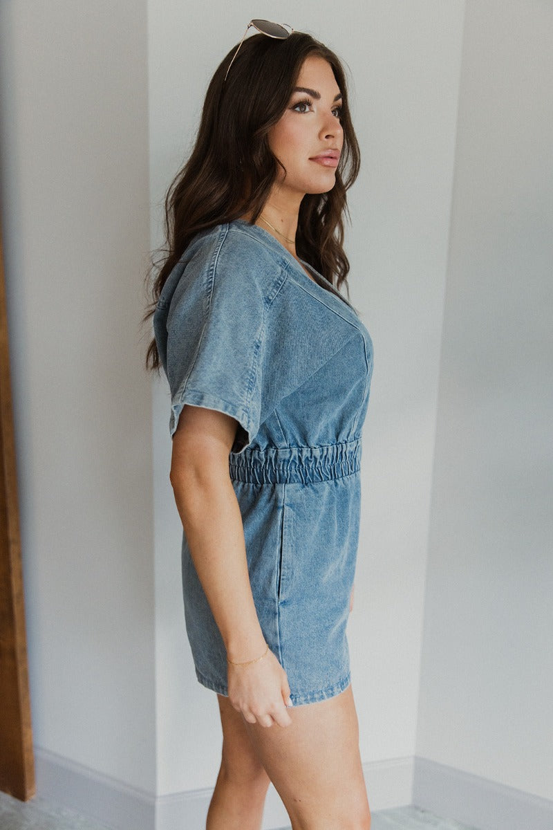 Side view of model wearing the Denim Dreams Romper that has light wash denim fabric, two front pockets, an elastic waistband, a front zipper closure, a plunge neckline and short sleeves