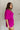 Side view of model wearing the Celia Romper in Magenta which features magenta lightweight fabric, a bubble hem, magenta lining, a tie around the waist, a surplice neckline, half puff sleeves with elastic trim, and a monochromatic back zipper with a hook c