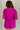 Back view of model wearing the Celia Romper in Magenta which features magenta lightweight fabric, a bubble hem, magenta lining, a tie around the waist, a surplice neckline, half puff sleeves with elastic trim, and a monochromatic back zipper with a hook c