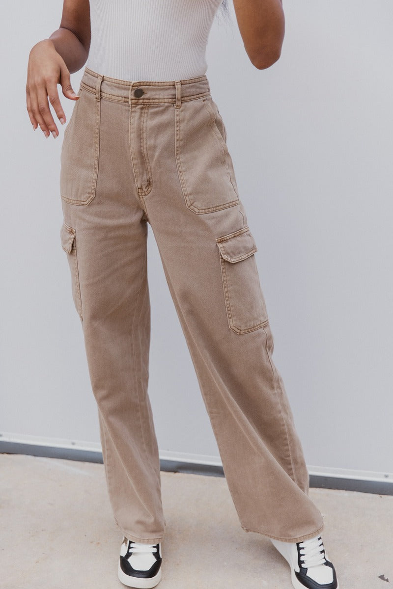 front view of model wearing the Ready or Not Cargo Pants in Brown that have light brown denim fabric, cargo pockets on each side, a front zipper with a button closure, two front pockets, belt loops and wide pant legs