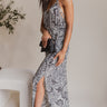 Full side view of model wearing the All I Want Maxi Dress that has a cream and black floral print, maxi length, a slit on the side, an elastic waist, a halter neckline with cutout details, adjustable straps, and an open back
