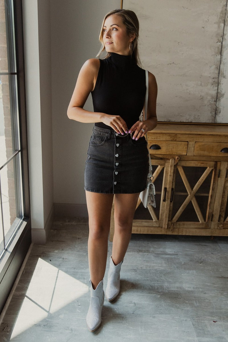 Full body front view of model wearing the Follow Your Heart Denim Skirt that has black denim fabric, mini length, two front pockets, two back pockets, belt loops, and white heart button-up closures