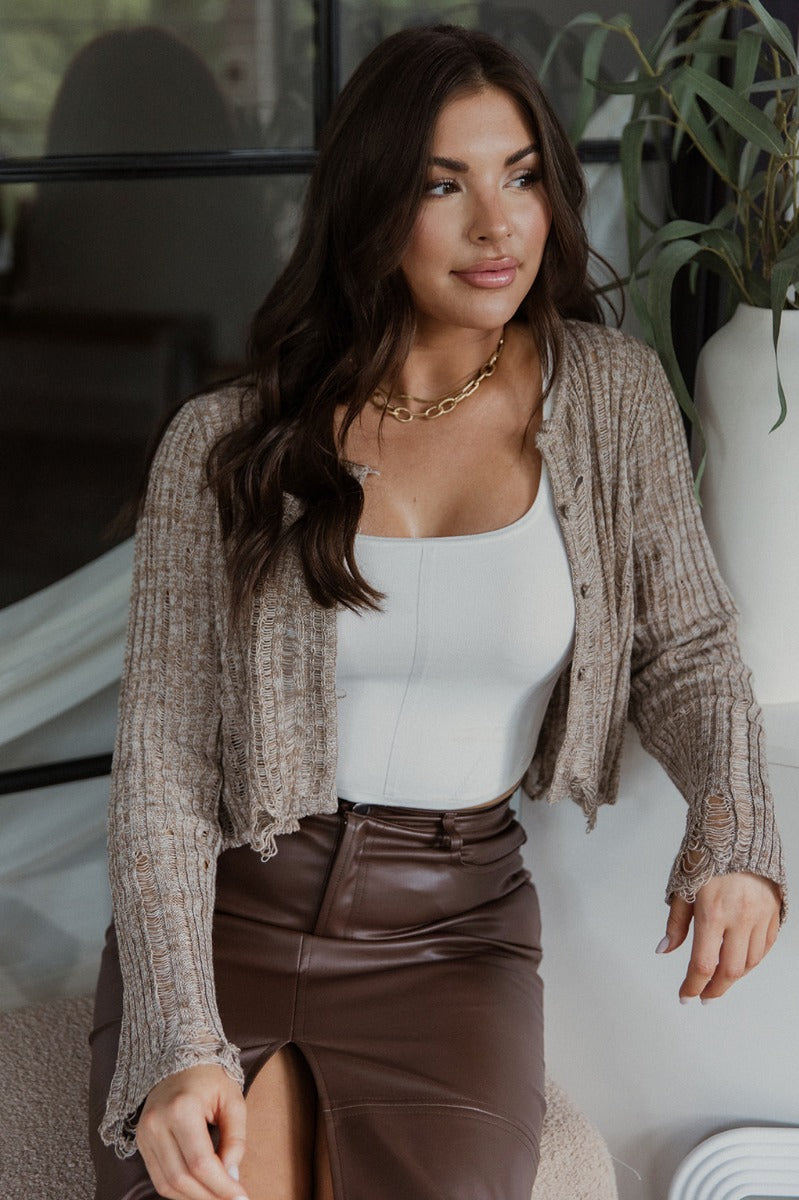 Front view of model wearing the Simply The Vibe Cardigan which features cream and brown knit fabric, distressed details, a cropped waist, covered button-up closures, a round neckline and long flare sleeves. The cardigan is open.