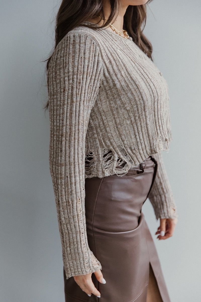 Side view of model wearing the Simply The Vibe Cardigan which features cream and brown knit fabric, distressed details, a cropped waist, covered button-up closures, a round neckline and long flare sleeves. The cardigan is closed.