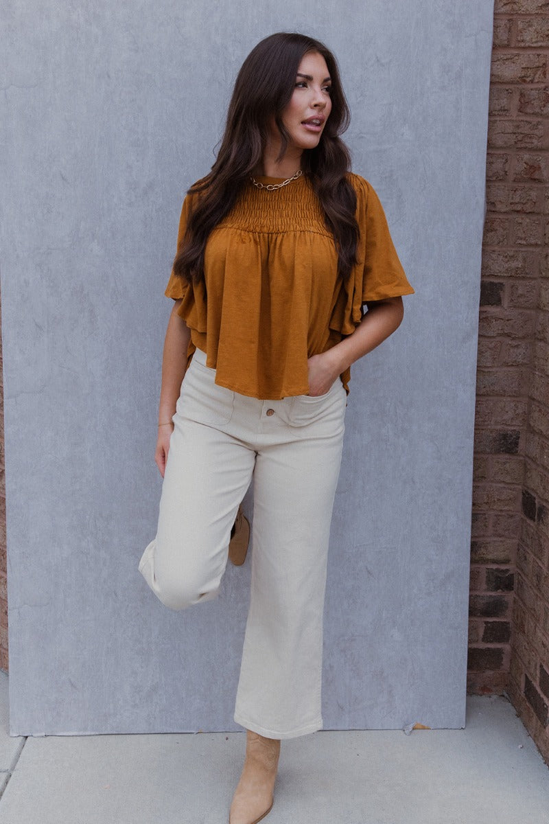 Full body view of model wearing the True Tradition Top which features camel knit cotton fabric, a scooped hem, a smocked upper, a round neckline and short flare sleeves.