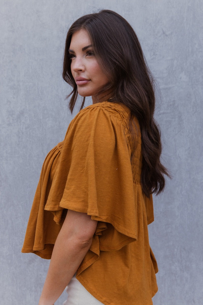 Side view of model wearing the True Tradition Top which features camel knit cotton fabric, a scooped hem, a smocked upper, a round neckline and short flare sleeves.