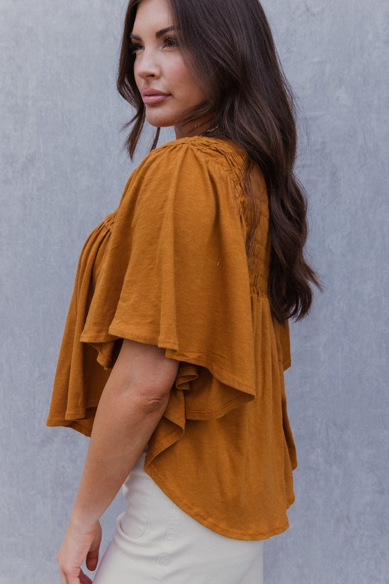 Side view of model wearing the True Tradition Top which features camel knit cotton fabric, a scooped hem, a smocked upper, a round neckline and short flare sleeves.