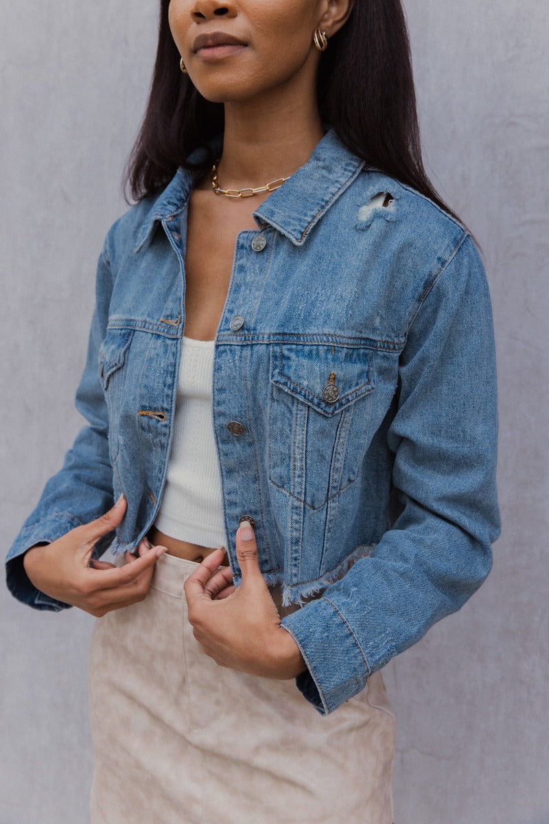 Close up view of model wearing the Wish You Were Here Denim Jacket which features washed denim fabric, a cropped waist, distressed details, button up closures, two front buttoned pockets, a collared neckline and long sleeve with buttoned cuffs.