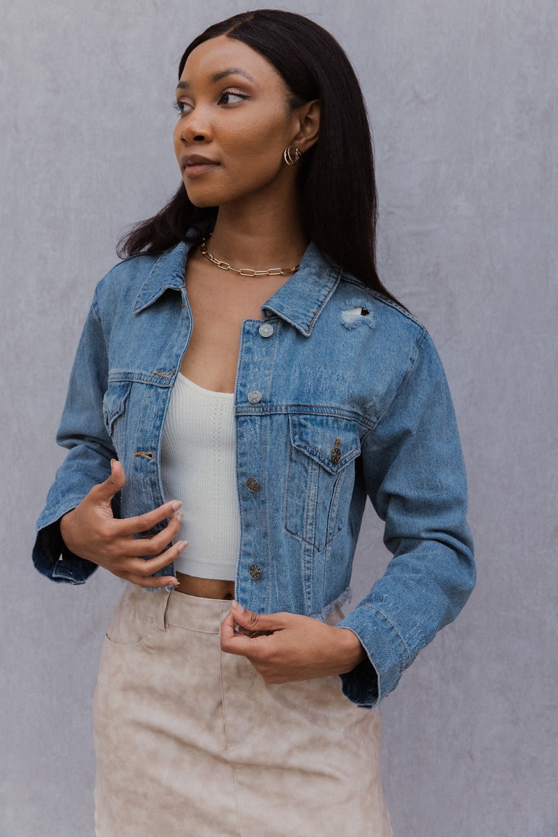 Frontal side view of model wearing the Wish You Were Here Denim Jacket which features washed denim fabric, a cropped waist, distressed details, button up closures, two front buttoned pockets, a collared neckline and long sleeve with buttoned cuffs.