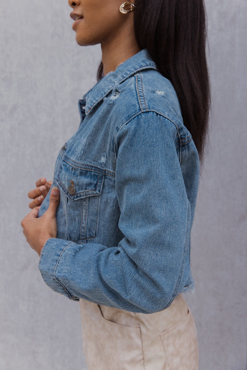 Side view of model wearing the Wish You Were Here Denim Jacket which features washed denim fabric, a cropped waist, distressed details, button up closures, two front buttoned pockets, a collared neckline and long sleeve with buttoned cuffs.