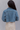 Back view of model wearing the Wish You Were Here Denim Jacket which features washed denim fabric, a cropped waist, distressed details, button up closures, two front buttoned pockets, a collared neckline and long sleeve with buttoned cuffs.
