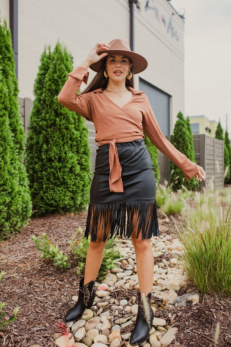 Full body front view of model wearing the Backstage Pass Skirt features a black faux-leather material, a thick elastic waist band, a high-waisted fit, fringe detailing along the bottom hem, and a mini length. Worn with brown top.