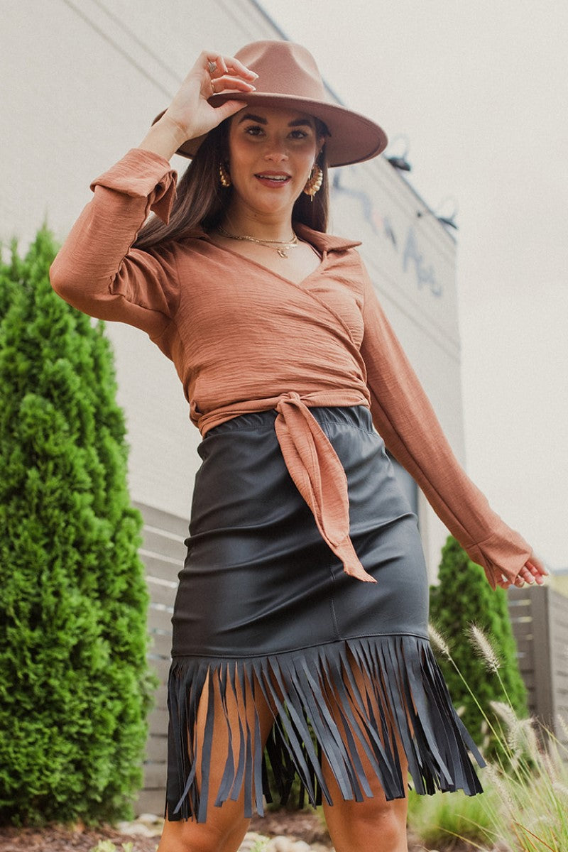 Front view of model wearing the Backstage Pass Skirt features a black faux-leather material, a thick elastic waist band, a high-waisted fit, fringe detailing along the bottom hem, and a mini length. Worn with brown top.
