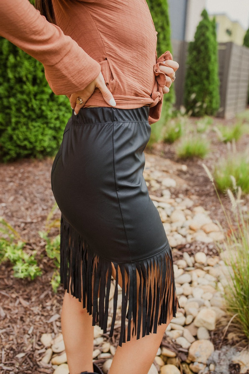 Close-up side view of model wearing the Backstage Pass Skirt features a black faux-leather material, a thick elastic waist band, a high-waisted fit, fringe detailing along the bottom hem, and a mini length. Worn with brown top.