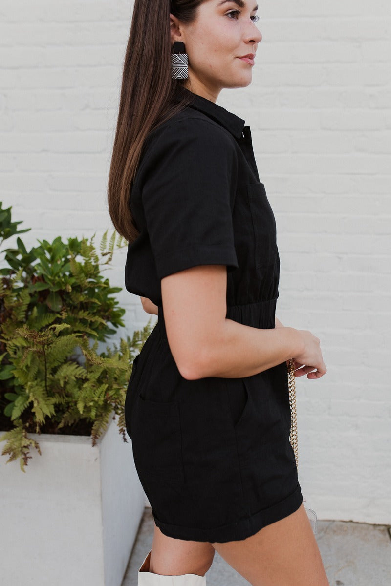 Side view of the Heading South Romper that features a black colored material, a collar neckline, a short cuffed sleeve, a button-up front, two front pockets at the chest, a cinched waist, two side pockets, cuffed bottom hem, and two back pockets.
