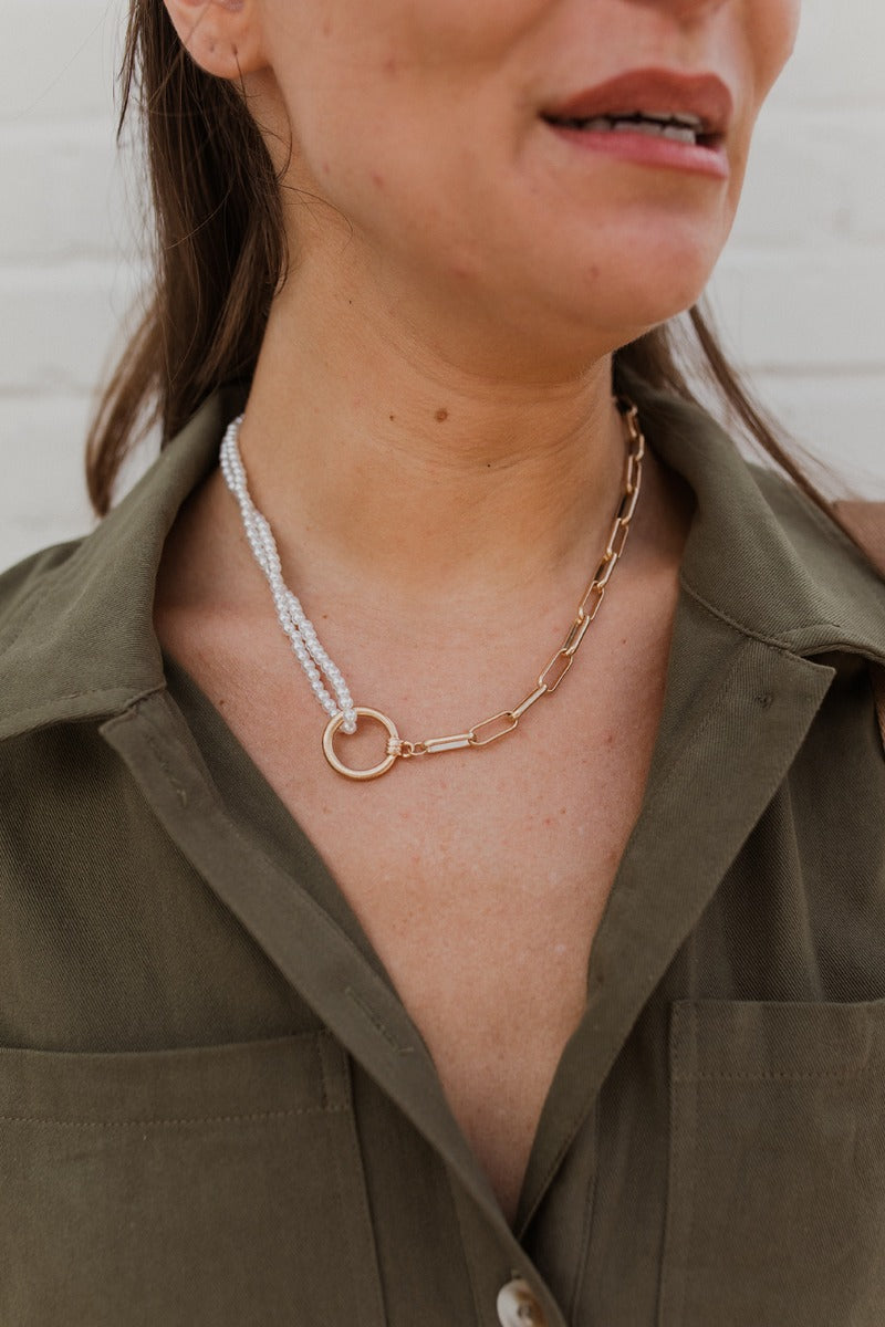 The About Time Necklace is a pearl necklace looped through a circle pendant with a gold chain link connected, finished with an adjustable link and clasp closure. 