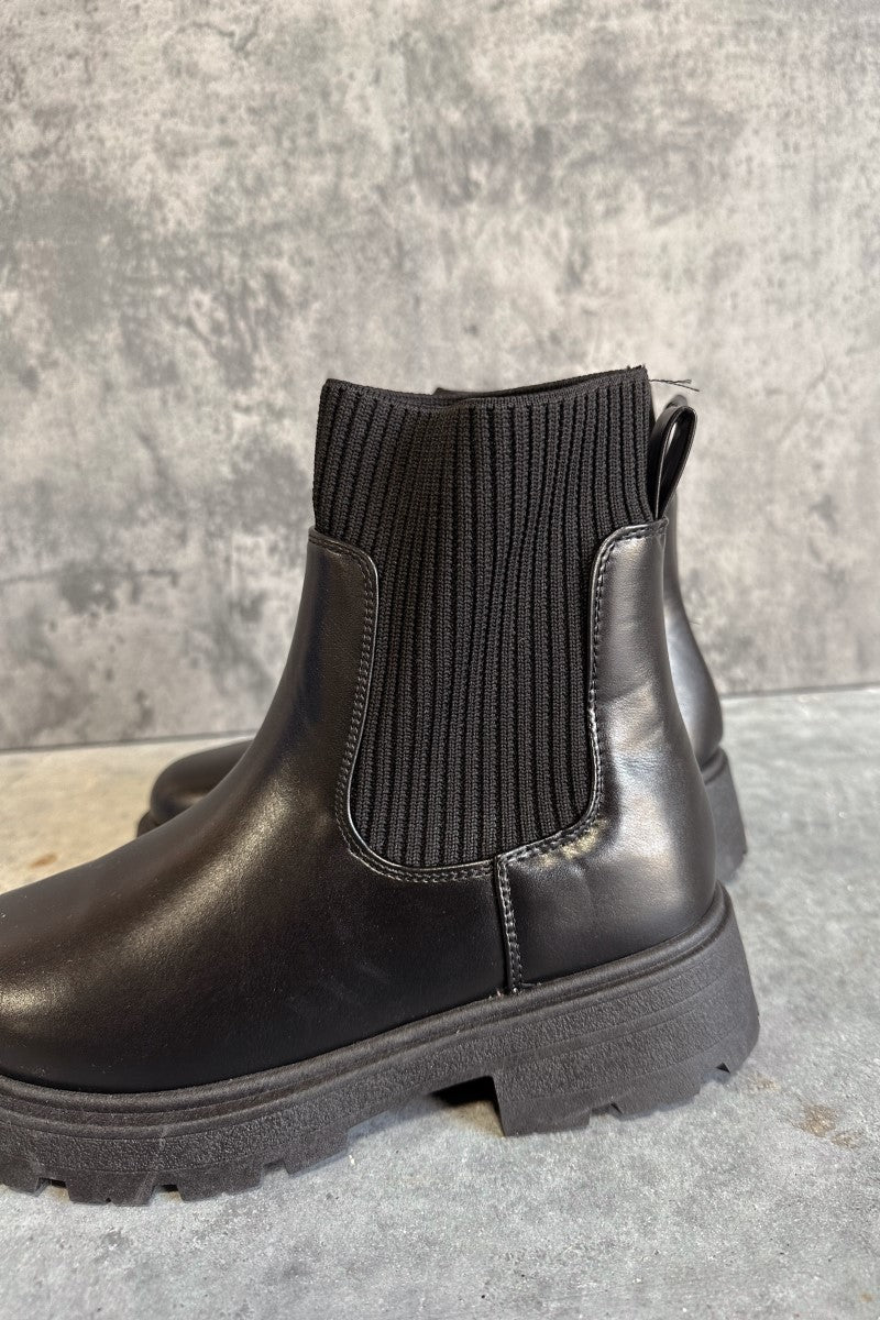 Side view of the Bronx Black Sock Booties that feature black faux-leather uppers with ribbed knit details against a grey background.