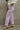 Side view of model wearing the Emily Lavender Wide Leg Pants which features acid lavender denim-like fabric, two front pockets, a front zipper with a button closure, belt loops, two back pockets, and wide legs.