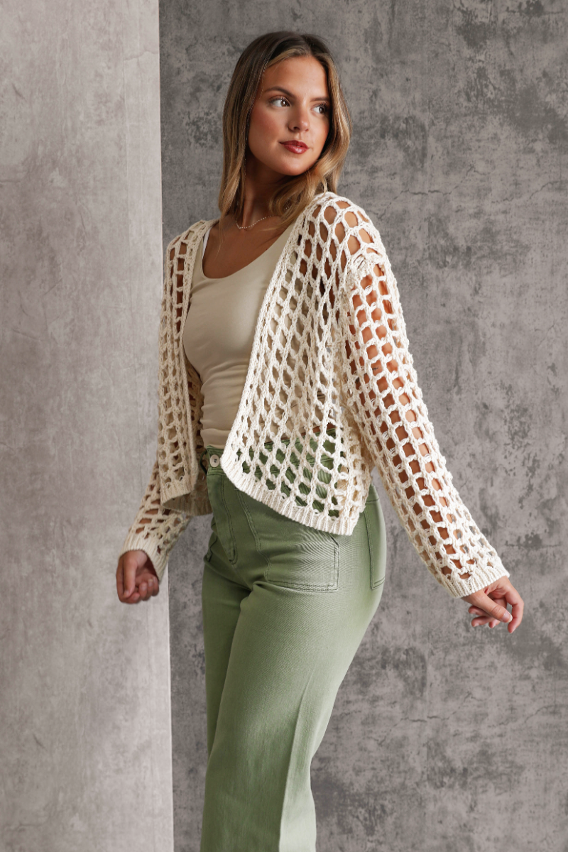 Side view of model wearing the Mila Cream Open Knit Cropped Cardigan which features cream open knit fabric, cropped waist, front opening with no closure.