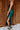 Side view of model wearing the Lainey Green Slit Midi Skirt which features teal and black shimmer pleated fabric, midi length, slit on the side, thigh length fuchsia lining and elastic waistband.