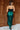 Back view of model wearing the Lainey Green Slit Midi Skirt which features teal and black shimmer pleated fabric, midi length, slit on the side, thigh length fuchsia lining and elastic waistband.