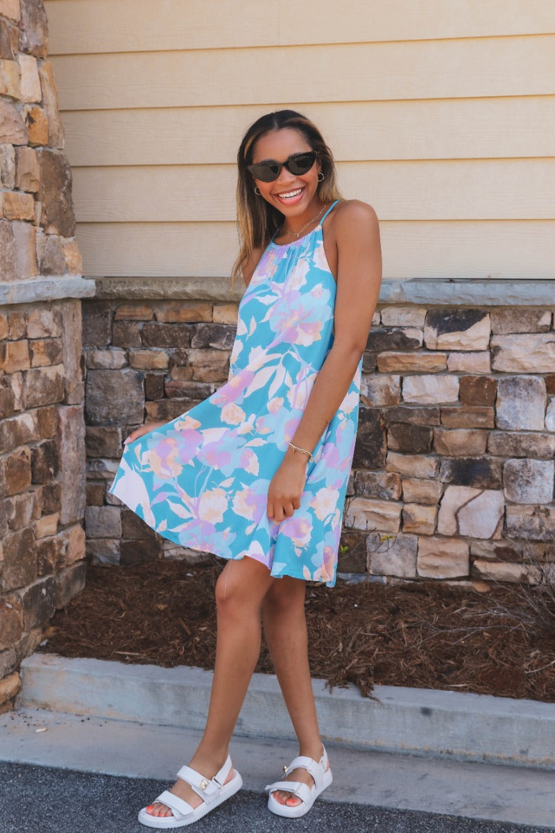 Full body front view of model wearing the Bahama Blues Floral Dress that has blue fabric with a pink, light blue, light pink and tan floral pattern, mini length, a halter neck, a sleeveless design, and a back keyhole with a tie closure.