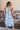 Back view of model wearing the Bahama Blues Floral Dress that has blue fabric with a pink, light blue, light pink and tan floral pattern, mini length, a halter neck, a sleeveless design, and a back keyhole with a tie closure.