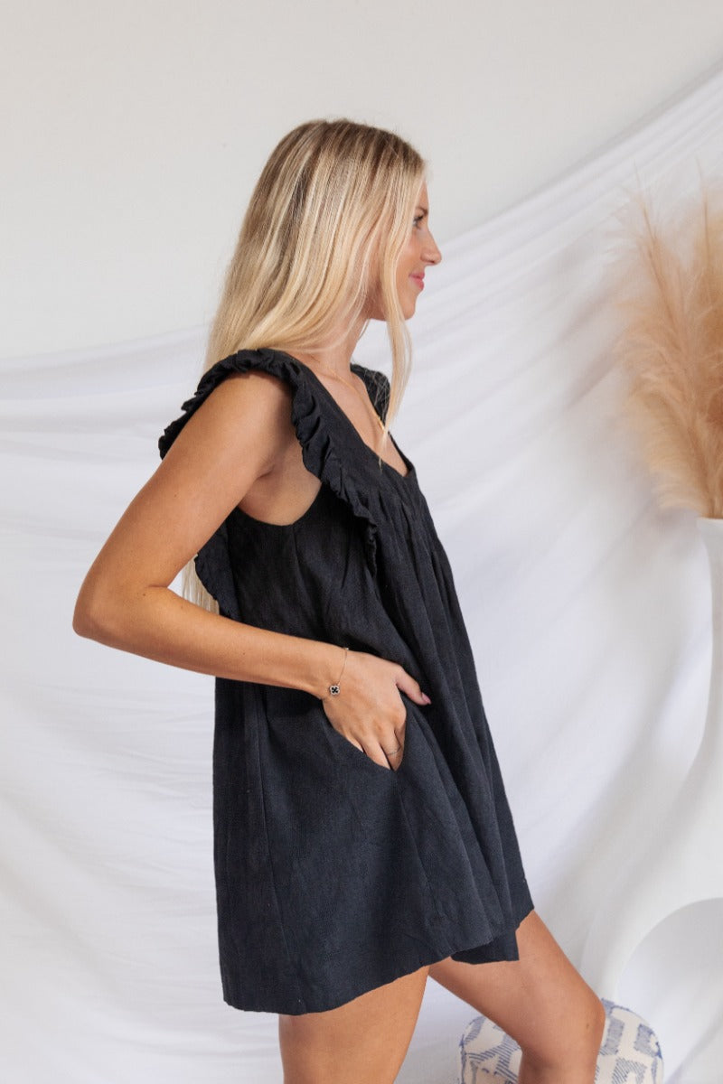 Side view of the End Game Romper that features a black cotton material, a rounded v neckline, a sleeveless design with tick straps, ruffle details at the straps, side pockets, a shorts bottom, a tie back, and a flowy fit.