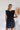 Back view of the End Game Romper that features a black cotton material, a rounded v neckline, a sleeveless design with tick straps, ruffle details at the straps, side pockets, a shorts bottom, a tie back, and a flowy fit.