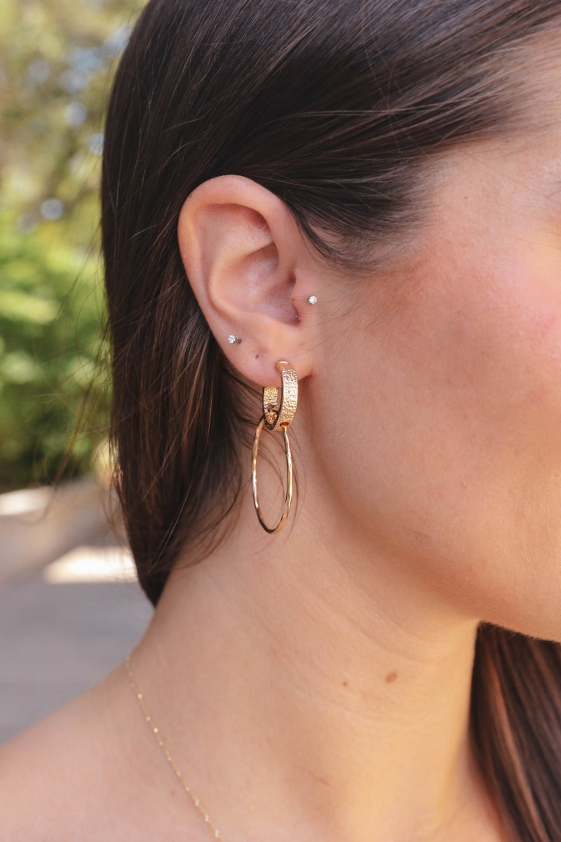Side view of model wearing the Attached At The Hoop Earring which features closed medium size gold hoops linked with an open mini, textured gold hoop.