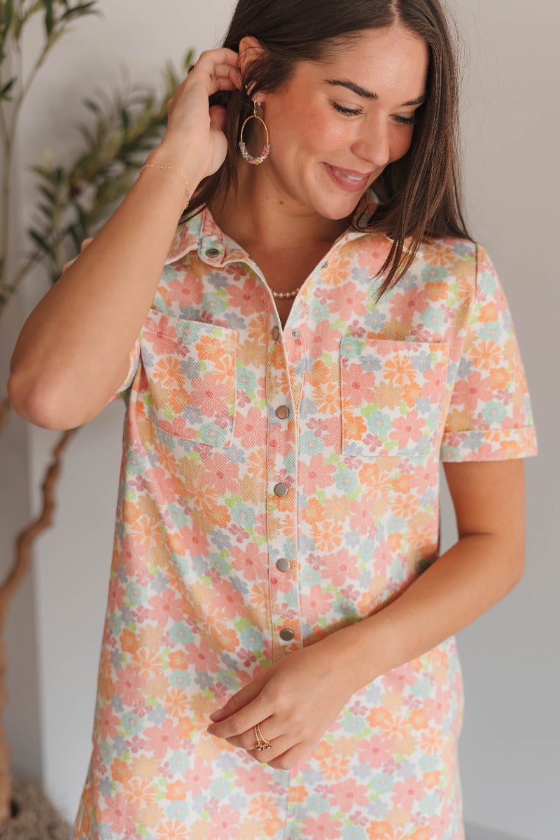 Upper front view of model wearing the Laguna Floral Romper that has denim fabric with an orange, pink, aqua, green and blue floral print, folded hem, two front chest pockets, side and back pockets, snap up buttons, a collared neck, and short sleeves