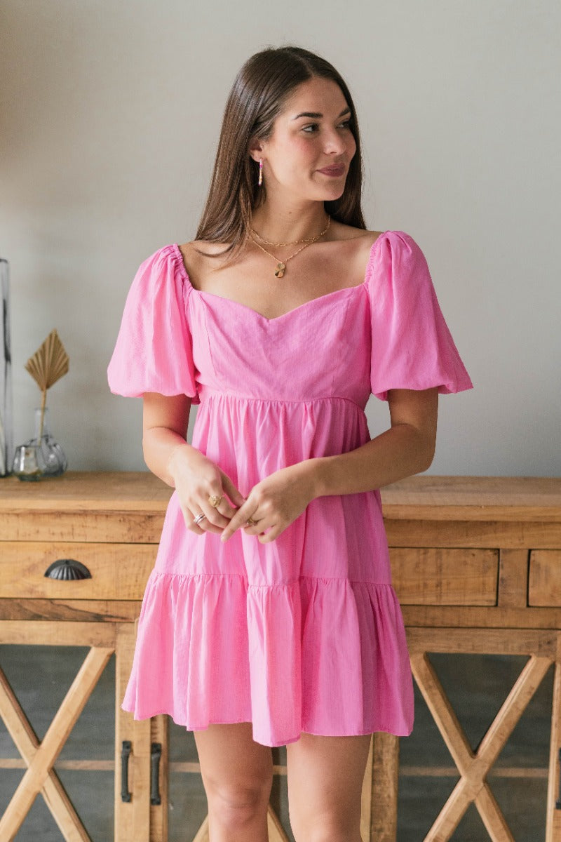 Front view of model wearing the Beautiful and Free Dress which features pink fabric, a flared peplum skirt, a square neckline, short puff sleeves with elastic trim, and an open back with a bow tie closure.