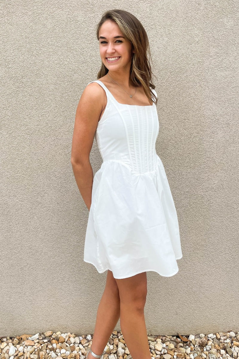 Front view of model wearing the Prepare To Swoon Dress which features white fabric, mini length, white lining, pockets on each side, boning upper details, square neckline, thick straps, sleeveless and monochromatic back zipper with hook closure.