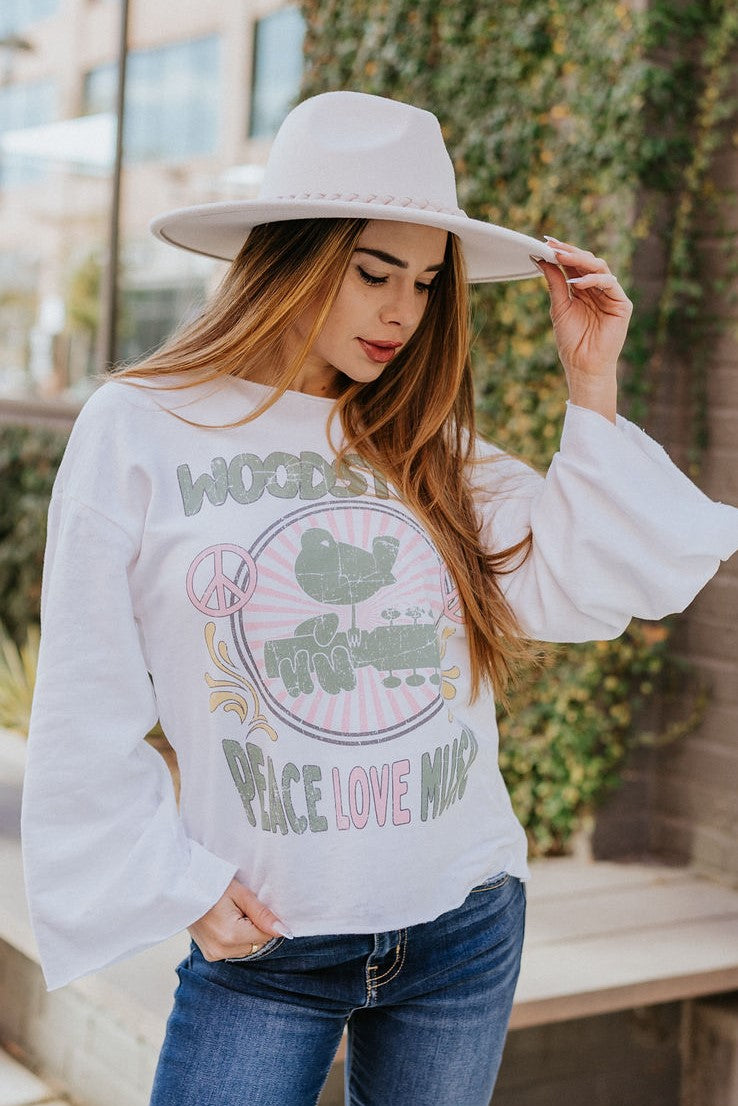 Front view of model wearing the Woodstock Graphic Top that has white fabric with a fleece interior, a green, pink and yellow graphic, a round, raw trim, and long bell sleeves. "Woodstock" and "Peace Love Music" displayed on the graphic