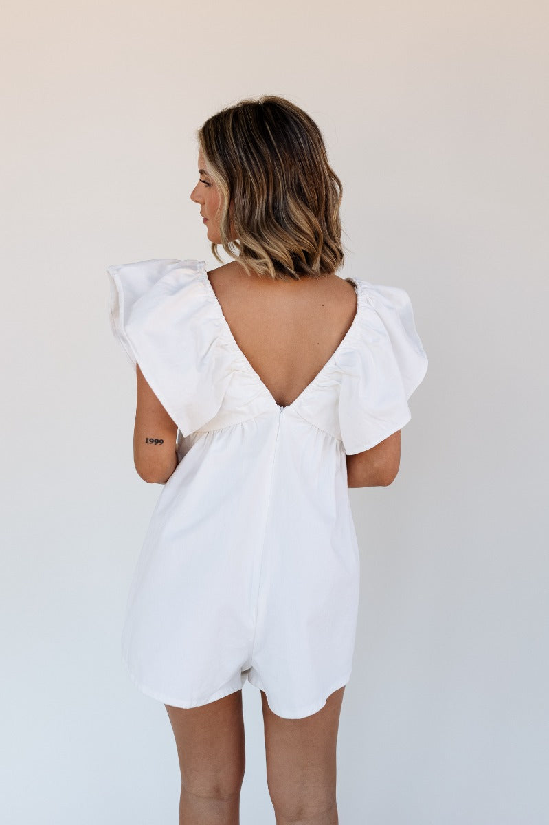 Back view of model wearing the Peyton Off White Denim Sleeveless Romper that has off white cotton fabric, two front pockets, a plunge neck, ruffle straps, and a back zipper with a hook closure.