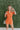 Frontal view of the Spur The Moment Denim Romper that features an orange denim material, a surplice neckline that snaps with a collar, half-length sleeves, a front zipper, pockets, pleating on the legs, and a 3" inseam.