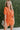 Frontal view of the Spur The Moment Denim Romper that features an orange denim material, a surplice neckline that snaps with a collar, half-length sleeves, a front zipper, pockets, pleating on the legs, and a 3" inseam.