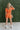 Back view of the Spur The Moment Denim Romper that features an orange denim material, a surplice neckline that snaps with a collar, half-length sleeves, a front zipper, pockets, pleating on the legs, and a 3" inseam.